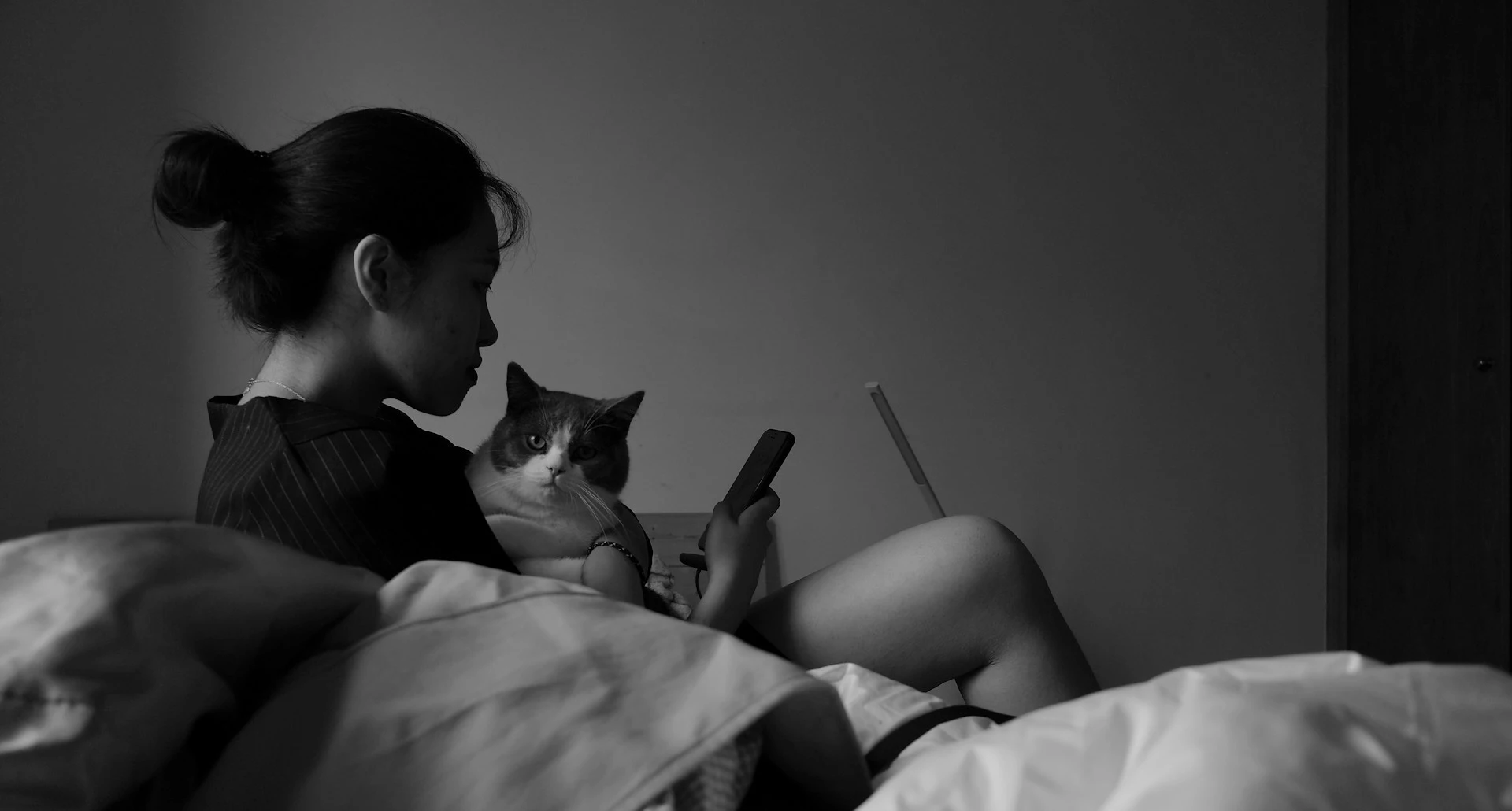 A black and white photo of a woman sitting on a bed, looking at her phone with a cat nestled on her lap.