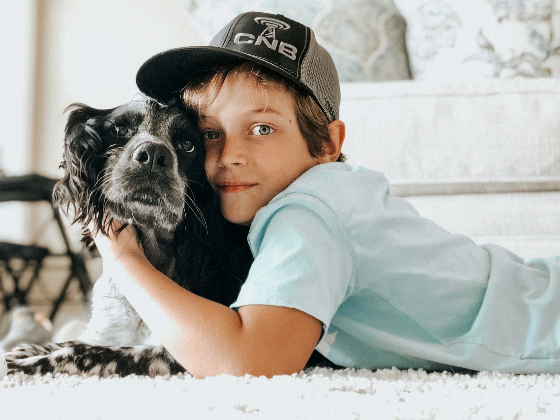 A boy is lying on a white rug, hugging a black and white dog.